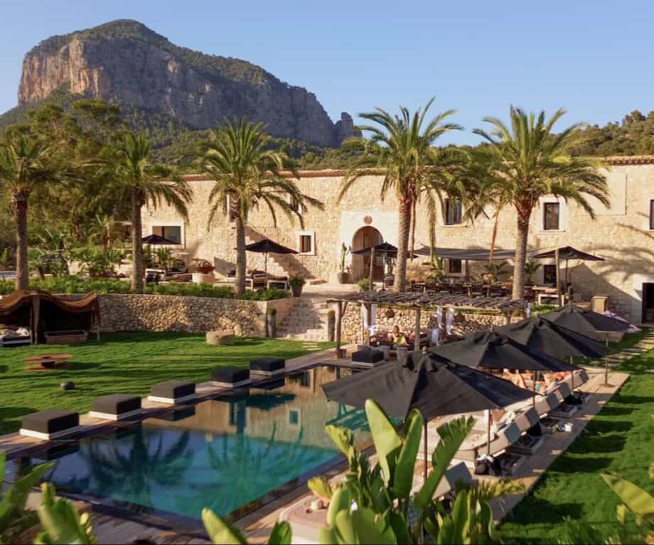 Luxury villa Son Fuster in Mallorca with palm-lined terraces, a large swimming pool, and outdoor lounge areas set against a dramatic mountain backdrop under clear blue skies.